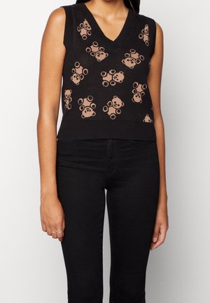 Gilet sans manches noir en coton avec un col en V et motif d'ours en peluche beige, porté avec un pantalon noir sur une personne debout.