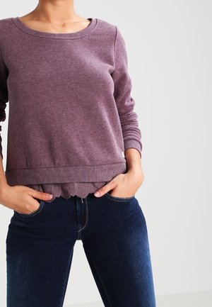 Vrouw draagt een mauve trui met lange mouwen over een top met golvende kanten rand, gecombineerd met donkerblauwe spijkerbroek, handen in de zakken.