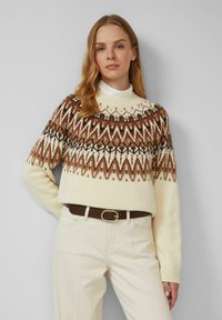 Gebreide trui in crème met een bruin geometrisch patroon op de schoudernaad, voorzien van een geribde col en lange mouwen, gecombineerd met lichte broeken.