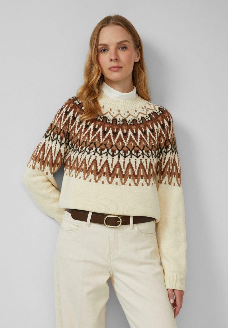 Gebreide trui in crème met een bruin geometrisch patroon op de schoudernaad, voorzien van een geribde col en lange mouwen, gecombineerd met lichte broeken.