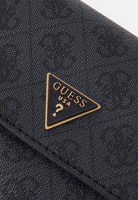 Musta teksturoitu lompakko, jossa on huomiota herättävä kultainen kolmikulmainen logo, jossa lukee "GUESS USA" ja kysymysmerkki. Materiaali on synteettinen, ja siinä on hienovarainen kuvio.