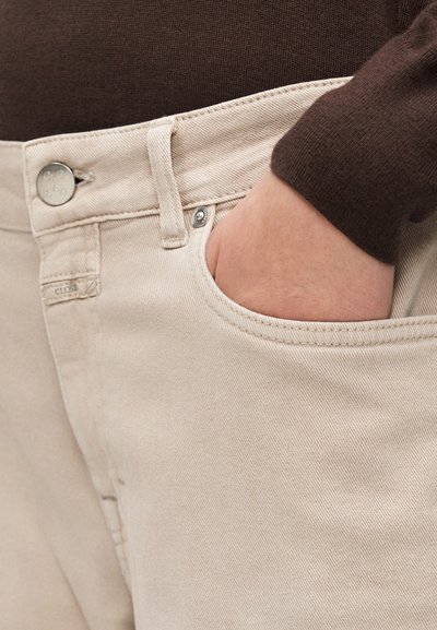 Main portant un pull à manches longues marron rentré dans la poche avant d'un pantalon beige avec rivets métalliques et fermeture à bouton.