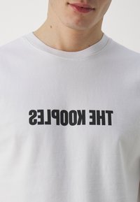 Bílé bavlněné tričko s výrazným černým invertovaným potiskem "THE KOOPLES". Kulatý výstřih, krátké rukávy, jemná textura.