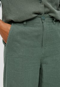 Lehké, texturované zelené kalhoty se rovný střih, předními kapsami a knoflíkovým zapínáním. Vyrobené z prodyšného materiálu podobného lnu.