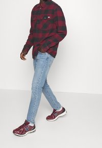 Camicia in flanella a quadri rossi e neri con tasche con bottoni, jeans aderenti blu chiaro e scarpe da ginnastica bordeaux con dettagli neri e bianchi.