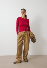 Rotes Langarmoberteil kombiniert mit weiten beigefarbenen Hosen. Der Stoff ist glatt und hat eine entspannte Passform. Das Model hält eine passende Jacke.