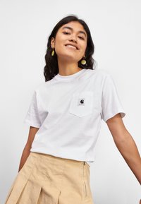 Hvit bomull T-skjorte med korte ermer, med en brystlomme og en liten svart logo. Modellen har på seg et plissert beige skjørt.
