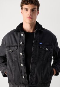 Chaqueta de mezclilla negra con un cuello peludo, cierre delantero de botones y dos bolsillos en el pecho; cuenta con herrajes plateados y un pequeño parche de logo azul.
