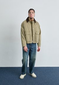 Khaki canvas jas met een bruin kraag, twee voorzakken, gedragen over een groene gebreide shirt, gecombineerd met blauwe spijkerbroeken en witte sneakers.