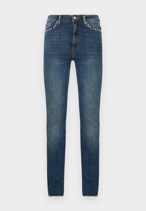 Jean skinny bleu avec des détails cloutés sur les poches avant et les passants de ceinture, et ourlet brut aux chevilles.