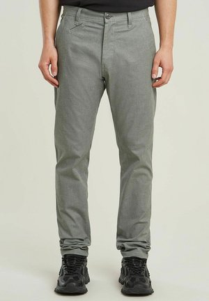 Homme portant un pantalon slim gris clair et des chaussures de sport noires, debout devant un fond uni.