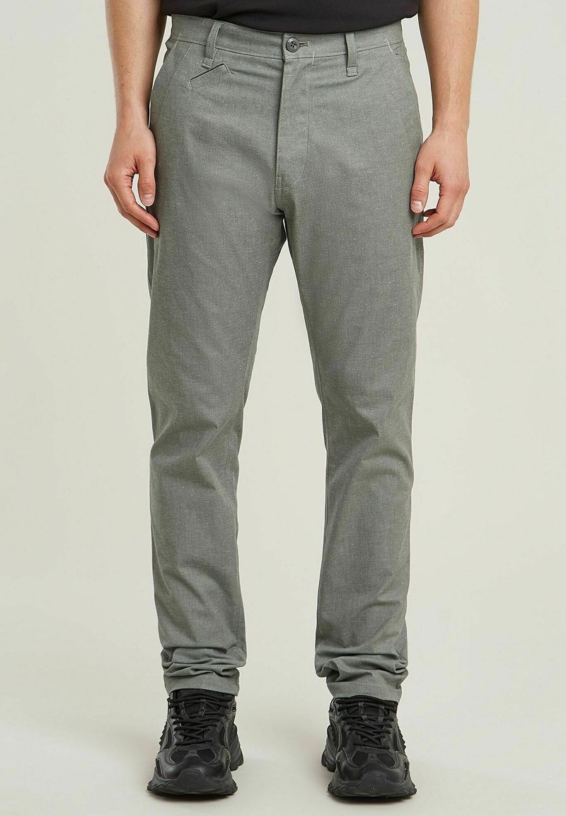 Homme portant un pantalon slim gris clair et des chaussures de sport noires, debout devant un fond uni.
