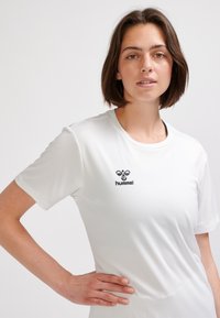 Biała sportowa koszulka z krótkim rękawem wykonana z lekkiego materiału, z okrągłym dekoltem i czarnym logo "hummel" po lewej górnej stronie.