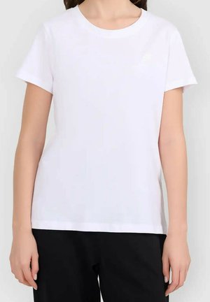 Kobieta nosząca gładką białą koszulkę z krótkim rękawem i czarne spodnie, pokazana od ramion do bioder na białym tle.