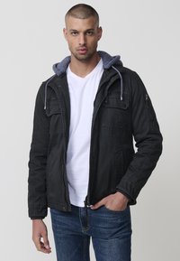 Melnā līkota jaka ar pelēku kapuci, ar divām priekšējām kabatām un kabatu uz rokām ar rāvējslēdzēju. Uzvilkta virs baltas T-kreklu un ziliem džinsiem.