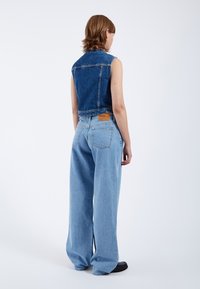 Chaleco de mezclilla con un diseño corto, combinado con jeans de tiro alto, sueltos y de color azul claro, que cuentan con un parche de cuero y bolsillos traseros. Calzado negro.
