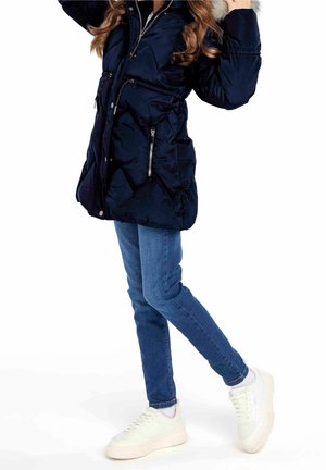 Enfant portant une veste d'hiver bleu marine, un jean bleu et des baskets blanches, se tenant debout avec une jambe pliée sur un fond blanc.