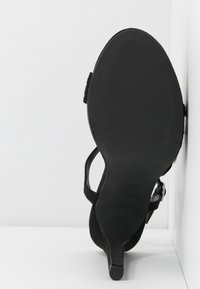 Sandale noire à talon avec la semelle tournée vers l'avant, posée contre un mur et un sol blancs en angle.