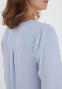 Blouse bleu clair avec une texture douce et lisse. Présente un design plissé le long du dos et des manches légèrement bouffantes.