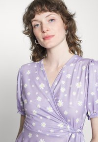 Vestido envelope roxo com estampa de margaridas brancas, mangas curtas e bufantes, e cintura atada. Tecido suave oferece uma textura lisa e um design casual.