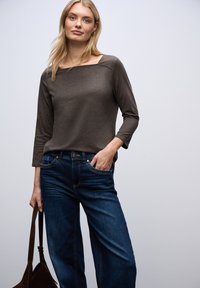 Tmavě hnědý dlouhý rukáv top s čtvercovým výstřihem, kombinovaný s modrými džínami. Modelka drží hnědou tašku a stojí na světlém pozadí.