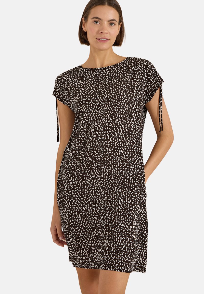 Femme portant une robe noire à manches courtes avec des points blancs abstraits et des accents noués aux manches, debout avec une main dans la poche.