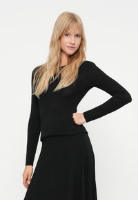 Νεαρή γυναίκα με μακριά ξανθά μαλλιά, φορώντας ένα μαύρο πουκάμισο με μακριά μανίκια και φούστα, πόζαρε με το ένα χέρι στη μέση μπροστά από έναν απλό λευκό τοίχο.