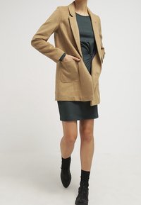 Blazer en mélange de laine beige avec une coupe ajustée, revers crantés et deux poches avant, porté sur une robe vert foncé. Des bottines noires à la cheville complètent le look.