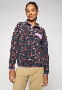 Fleece-pullover med flerfarget dyremønster i nyanser av brunt, teal og rosa, samt en kontrasterende rosa krage og lomme.