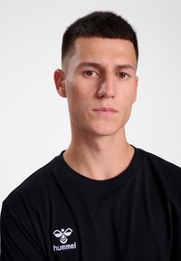 Zwart katoenen T-shirt met een ronde hals en een geborduurd wit logo. Model heeft kort haar en een neutrale uitdrukking.