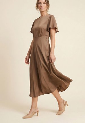 Vrouw die loopt in een vloeiende bruine midi-jurk met fladdermouwen en beige slingbacks met midhakken tegen een effen achtergrond.