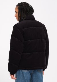Schwarze Cord-überwurfjacke mit hohem Kragen, gepolstertem Design und umgeschlagenen Ärmeln. Getragen mit hellblauen Jeans, die eine strukturierte Oberfläche zeigen.