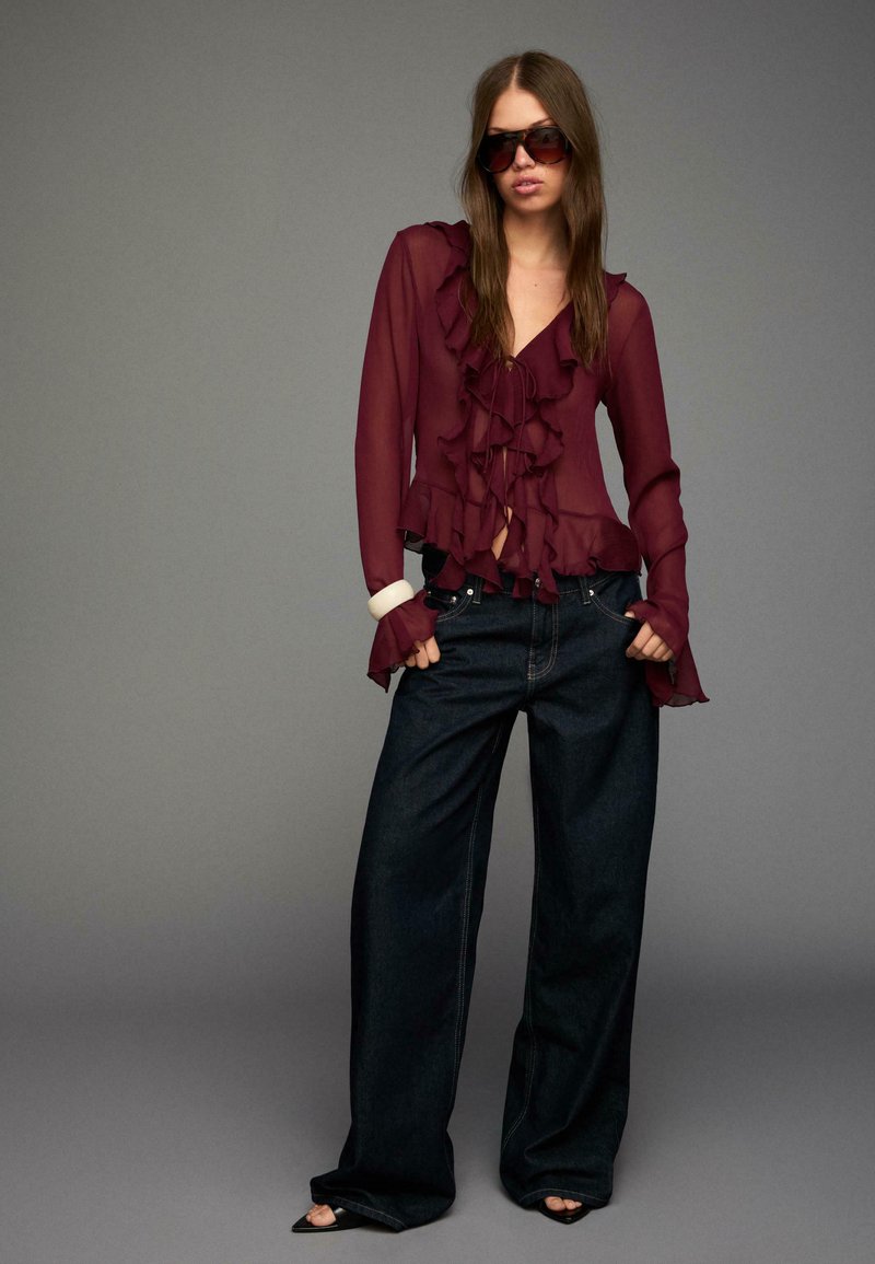 Burgundy chiffon blouse with ruffles, paired with loose dark denim jeans. Blouse features a v-neck and flared sleeves. Accessorized with sunglasses.