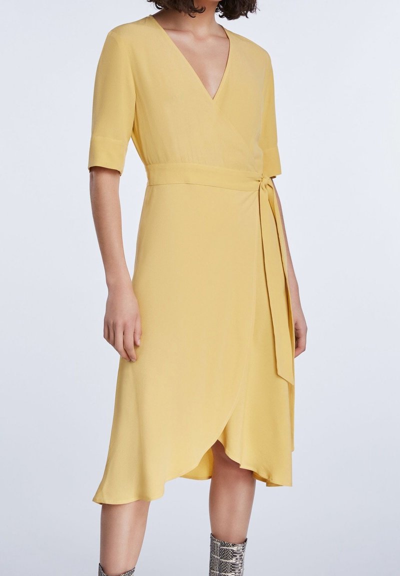 Gul omlottklänning med V-ringning, korta ärmar och knytning i midjan. Mjuk tyg med en svängd nederkant och diskreta detaljer.