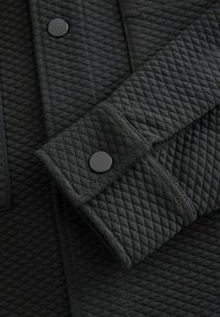 Schwarze gesteppte Jacke im Detail, die einen strukturierten Stoff zeigt, mit einer flachen Tasche und runden schwarzen Knopfakzenten am Ärmel.