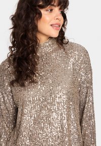 Top decorativo a maniche lunghe con scollo alto, caratterizzato da un motivo di paillettes scintillanti in tonalità dorate e argentate su una superficie testurizzata.