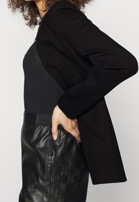 Černý přizpůsobený blazer s dlouhými rukávy, kombinovaný s upnutým černým topem a texturovanými černými kalhotami se vzory hvězd v zelených akcentních barvách.