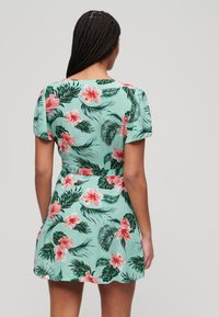 Bloemige jurk met korte mouwen, met een groene achtergrond, roze hibiscus bloemen en bladpatronen. Aansluitend bij de taille, wijd uitlopend aan de zoom.