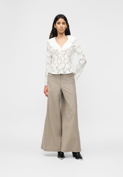 Γυναίκα με μακριά σκουρόχρωμα μαλλιά, ντυμένη με λευκό μπλουζάκι από δαντέλα, φαρδιά μπεζ παντελόνια και μαύρα μπότες με ψηλό τακούνι και μυτερή μύτη, μπροστά από ένα απλό φόντο.