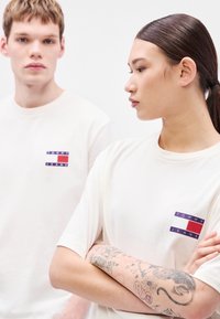 T-shirts de algodão cor creme com um pequeno logótipo retangular em azul-marinho, vermelho e branco no peito esquerdo. Mangas curtas, corte descontraído.