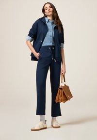 Sweat à capuche bleu marine sur une chemise en denim avec un pantalon bleu marine à cordon, des baskets blanches et un sac à main en cuir marron, avec des accents en métal argenté.