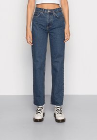 Högmidjade blå denimjeans med rak passform, med kontrastsömmar, bälteshällor och knappstängning. Bärs med vita snörstövlar.