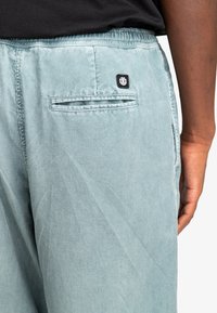Light blue corduroy pants with an elastic waistband, featuring a back pocket and a small black logo patch. Textured fabric with vertical lines.