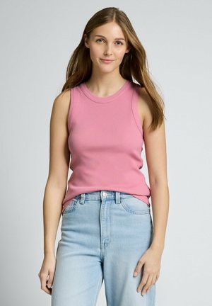 Giovane donna con lunghi capelli castani, che indossa un top rosa senza maniche e jeans a vita alta azzurri, in piedi davanti a uno sfondo grigio uniforme.