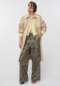 Beige színű ballonkabát visszahajtott ujjakkal, világos sárga kardigán fölött viselve. A bőszárú nadrág barna alapon zöld és kék absztrakt mintákkal díszített.