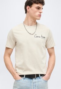 Νέος άντρας που φοράει μπεζ μπλούζα Calvin Klein, ασημένιο κολιέ αλυσίδας, ανοιχτό μπλε τζιν και μαύρη ζώνη, κοιτώντας προς τα αριστερά του.