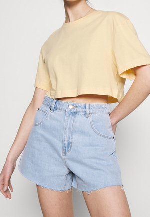 Persoon draagt een losse gele cropped t-shirt en lichtblauwe hoogwater spijkershorts met rafelige zoom tegen een effen achtergrond.