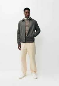 Graue Jacke mit texturiertem Design, getragen über einem Strickpullover, kombiniert mit beigen Hosen und weißen Sneakers. Schlichtes, modernes Erscheinungsbild.