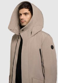 Värillinen parka, jossa on suuri huppu, vetoketjullinen sivutasku ja nappikoriste. Kangas näyttää sileältä ja sillä on matta pinta.