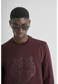 Sudadera burdeos con un gráfico de tigre oscuro y texturizado; escote redondo. Gafas de sol con montura transparente y lentes oscuros. Estilo sencillo y moderno.